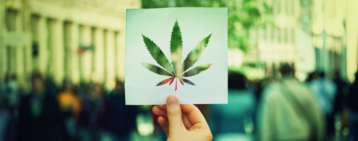 Hand, die ein Papierblatt mit einem Symbol für Marihuana-Blätter auf einem überfüllten Straßenhintergrund hält.
