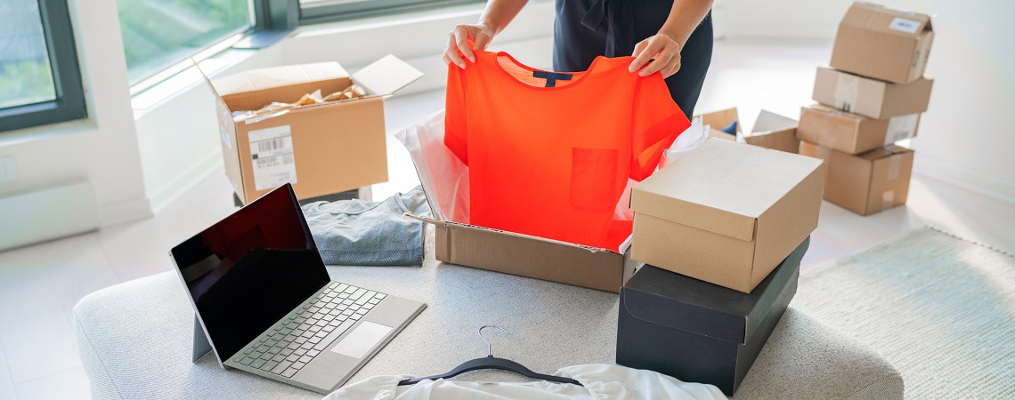 Retoure: Person zieht T-Shirt aus Paket neben Laptop und Paketen